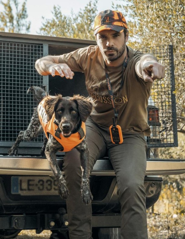 A man wearing a beige Percussion Compagnie T-shirt with wild boars on it, standing next to his car with his dog